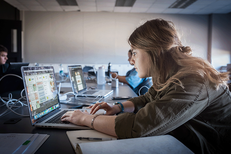 Student at a laptop in a computer lab with paper and pencil studying