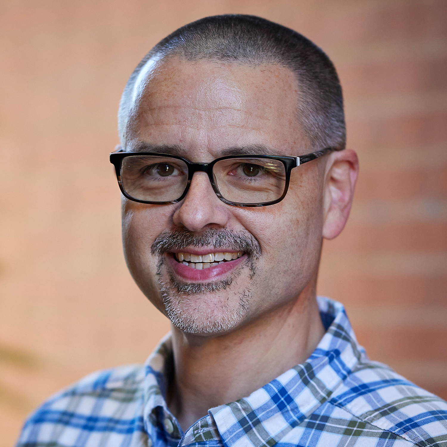 man in blue and white checked shirt wearing glasses with short cropped dark hair