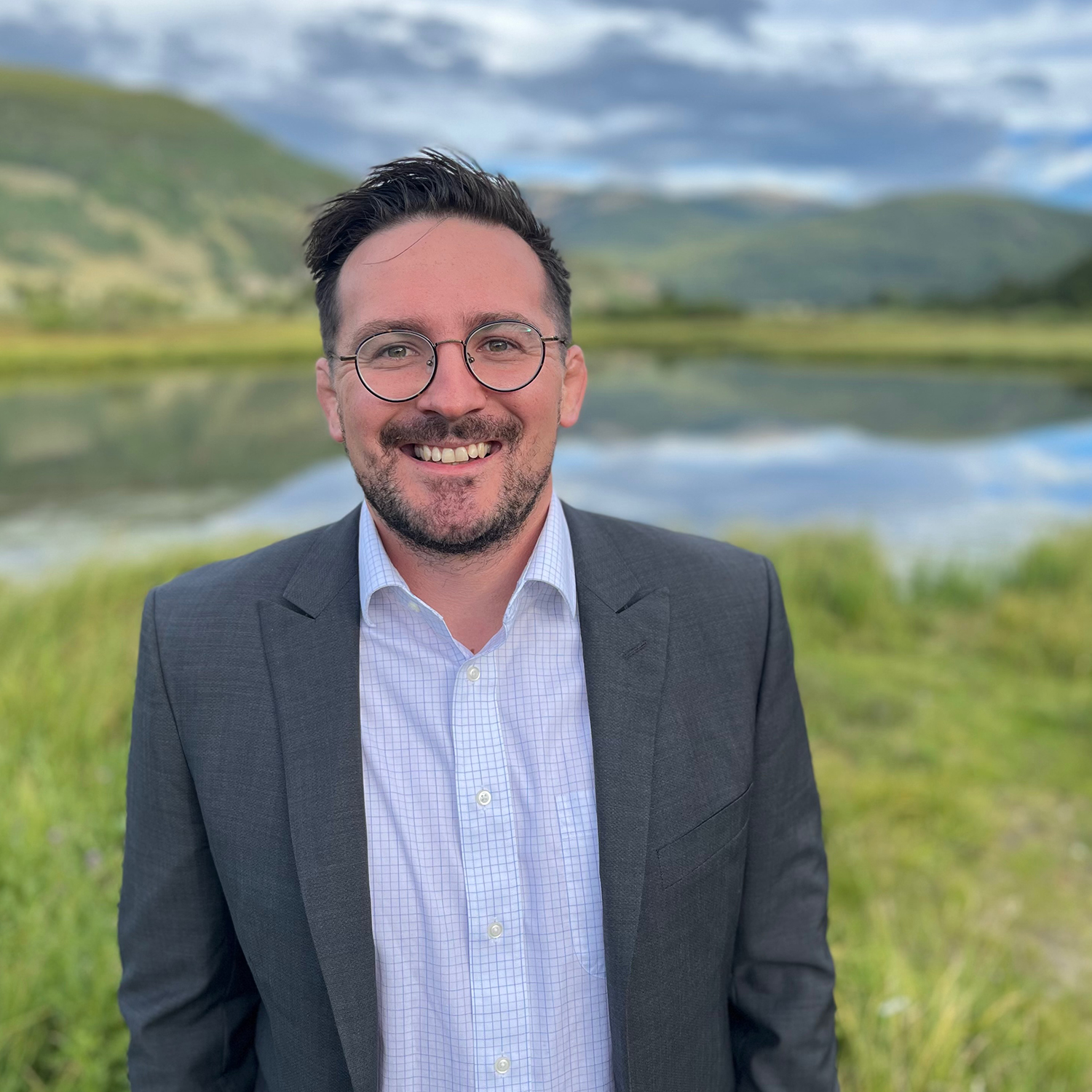 Corey Peltier in a light blue shirt and blue blazer standing in front of a lake