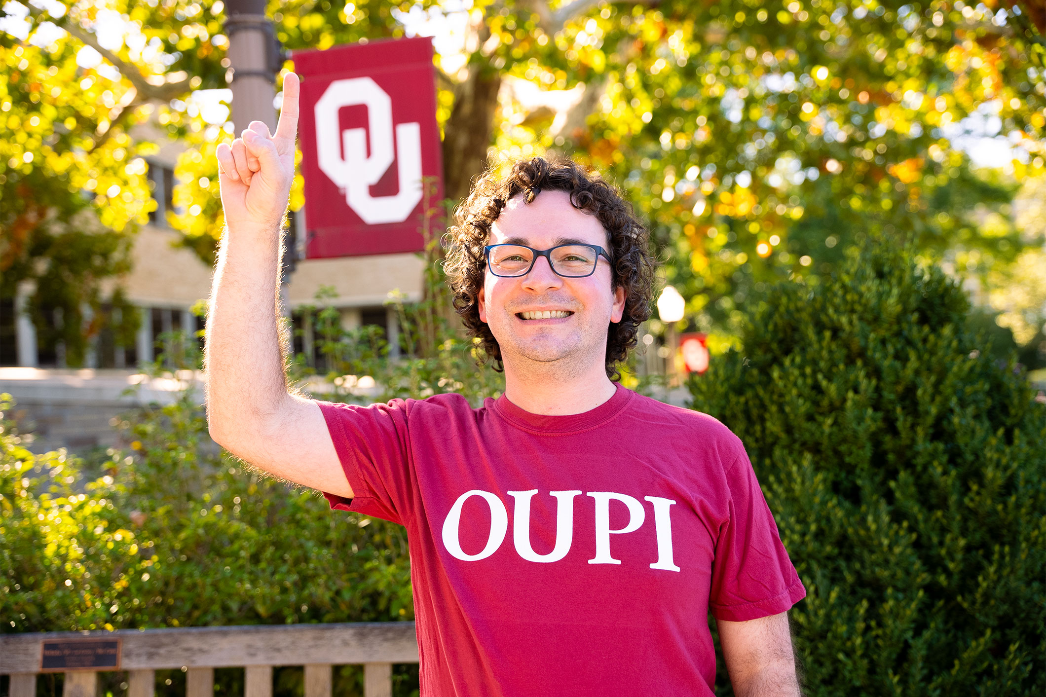 An individual wearing a crimson t-shirt that reads "OUPI" raises their index finger; in the background is an OU flag.