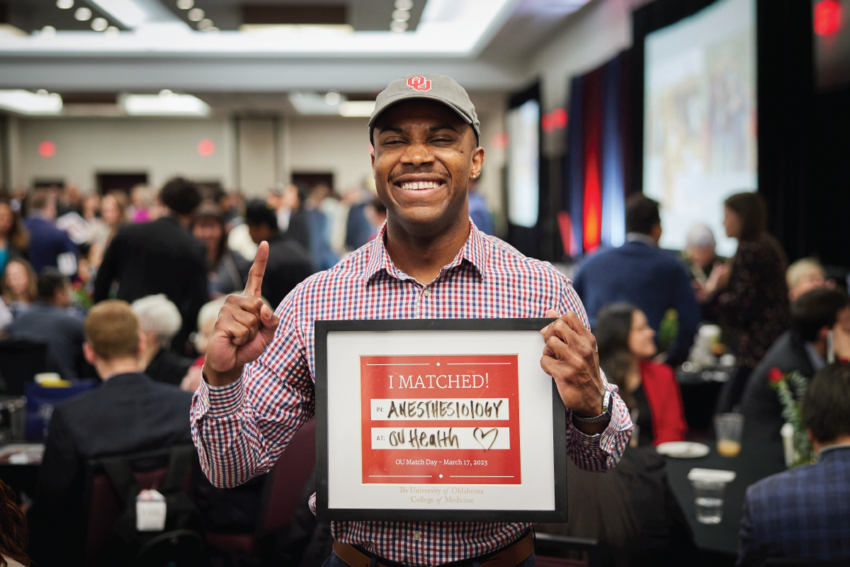 A smiling OU student wearing a baseball hat holds up their pointer finger and their Match Day assignment.