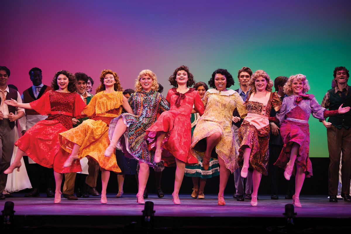 A line of students in costume, on stage, performing a dance.