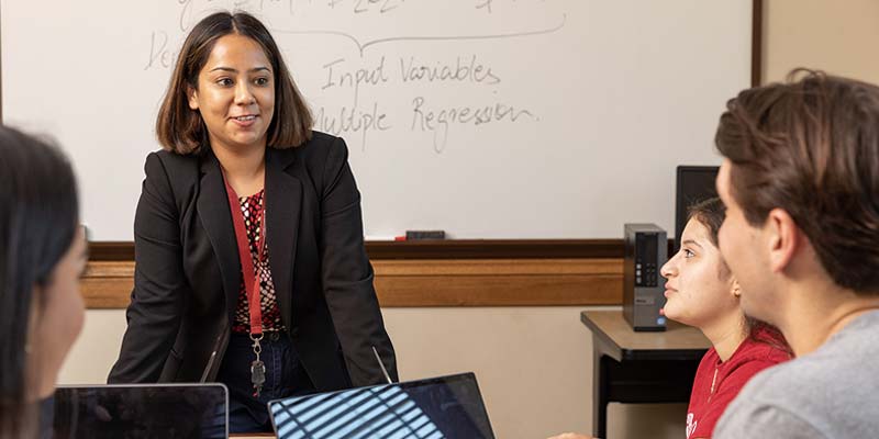 A professor standing at the front of the classroom in front of a white board, three students sit nearby. 