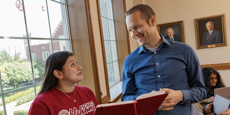 Professor and student look over a book together in a study room with large window that overlooks the Norman OU campus. 