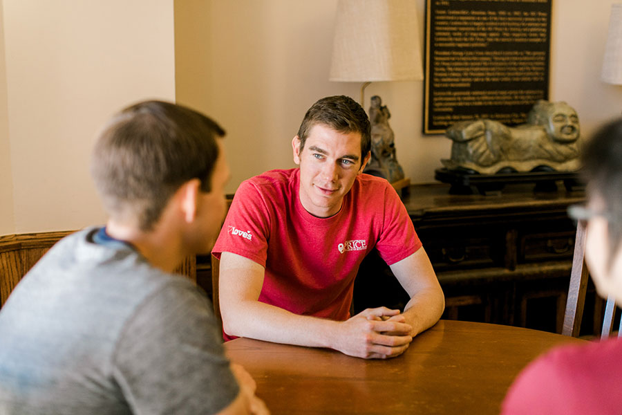 A student sits at a table while having a concersation with twho other people. 