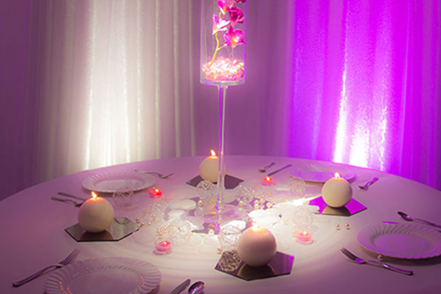 A round table dining setting with plates and utensils. Tall centerpiece in the middle with white light shining on it.