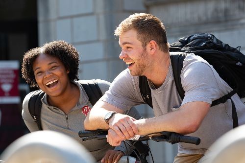 Two students stand outside near the the bike racks making conversation and laughing