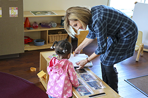 A woman takes notes while a teacher and children work on crafts projects at a table.