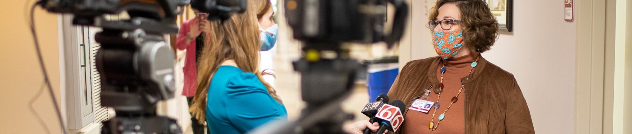 A reporter holding two microphones interviews a masked student on campus