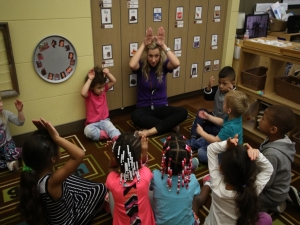 A teacher sits on a rug surrounded by students all mimicking her holding her hands over her head like bunny ears