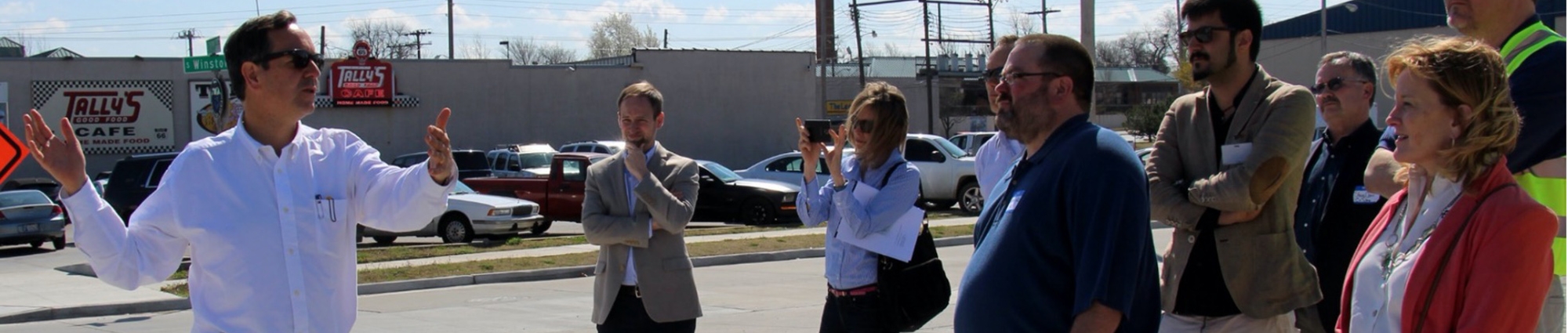 An urban planner gestures with both hands as he explains a city plan to a group of gathered students. A bright orange Road Work sign sits nearby