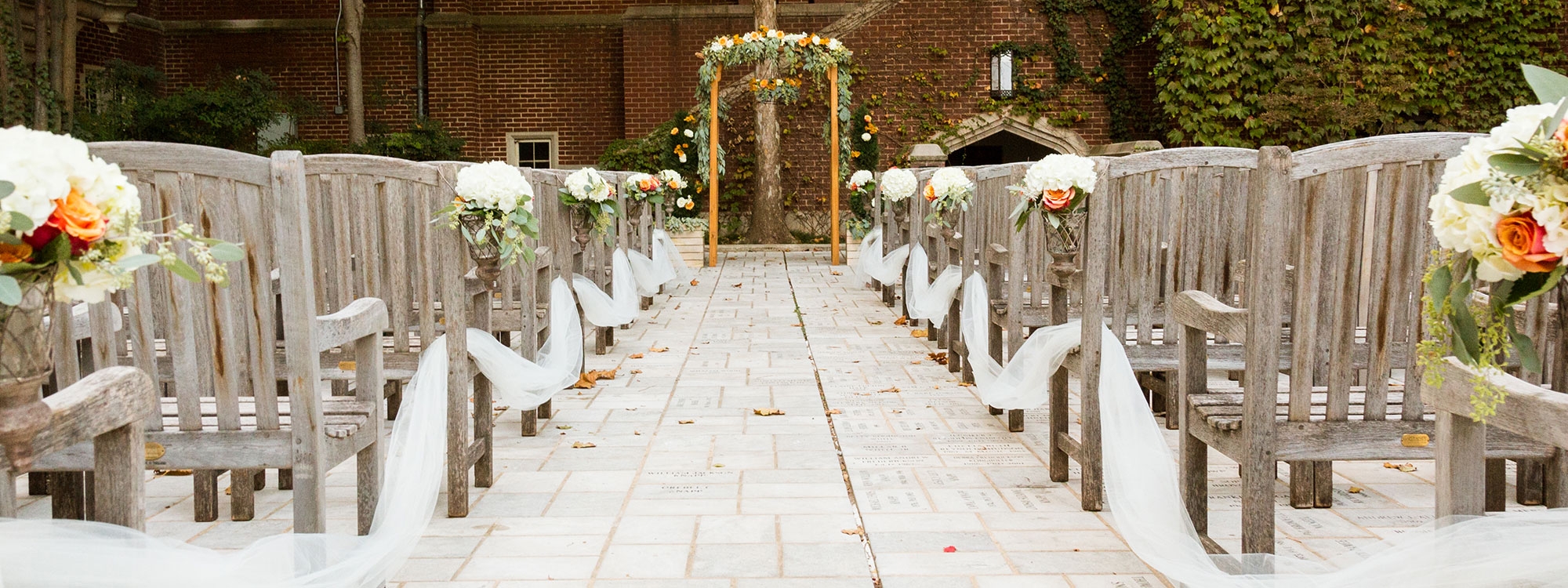 An aisle on stone pavers leading to a wooden arbor with flowers, wood chairs on sides of aisle with flowers.